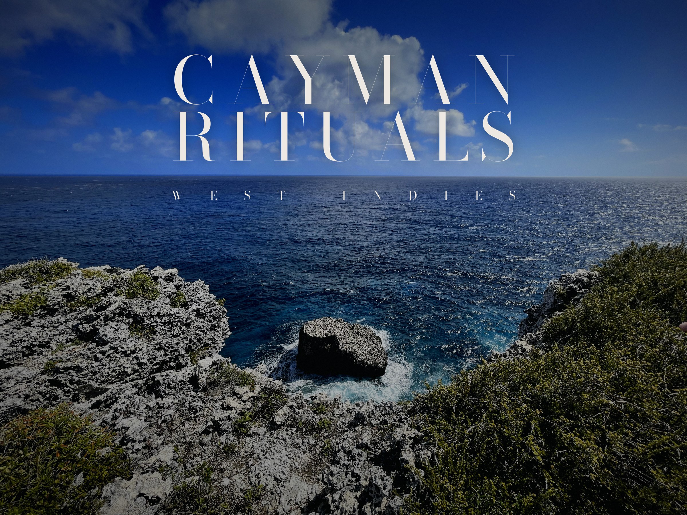 Looking out from the top of the Bluff on Cayman Brac: grey ironshore and low scrub in the foreground, a rock islet in white water directly below, and open blue sea to the horizon under a sky of small clouds.
